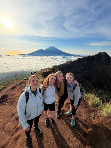 Four people standing on a mountain trail with a view of a volcano and clouds below. 4