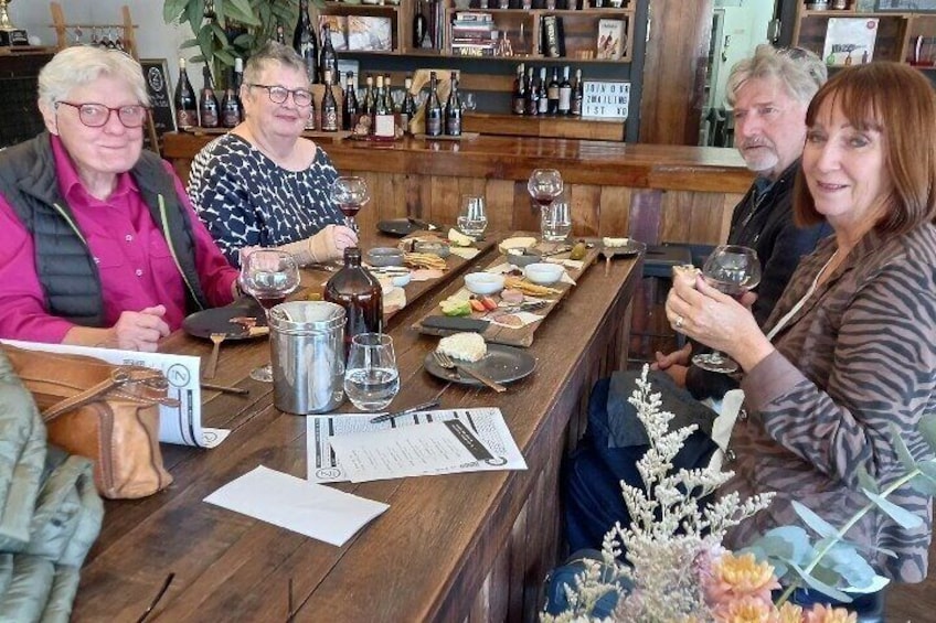 A group of people seated at a wooden table with wine glasses, bottles, and food items. 1