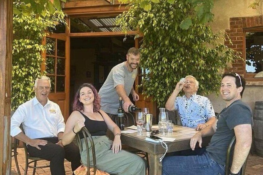A group of people sitting at an outdoor table with drinks and a bottle of wine, surrounded by greenery and a brick building. 5