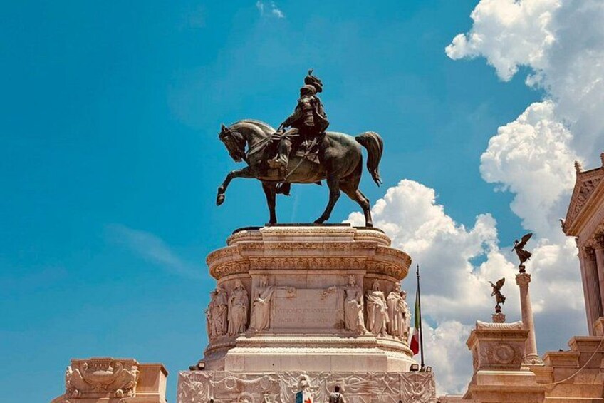 Une statue représentant une personne à cheval, posée sur un piédestal, avec un ciel bleu clair et des nuages en arrière-plan. 3
