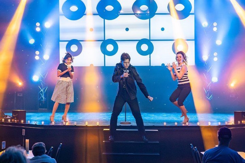 A stage with three performers, a large screen with abstract designs, and stage lighting. 3