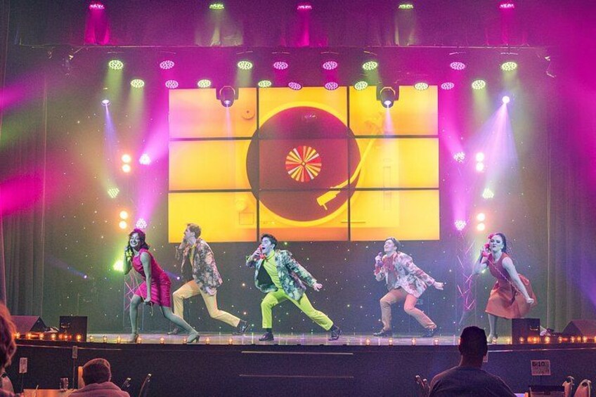 A stage with performers in colorful costumes, a large illuminated screen, and stage lights. 2
