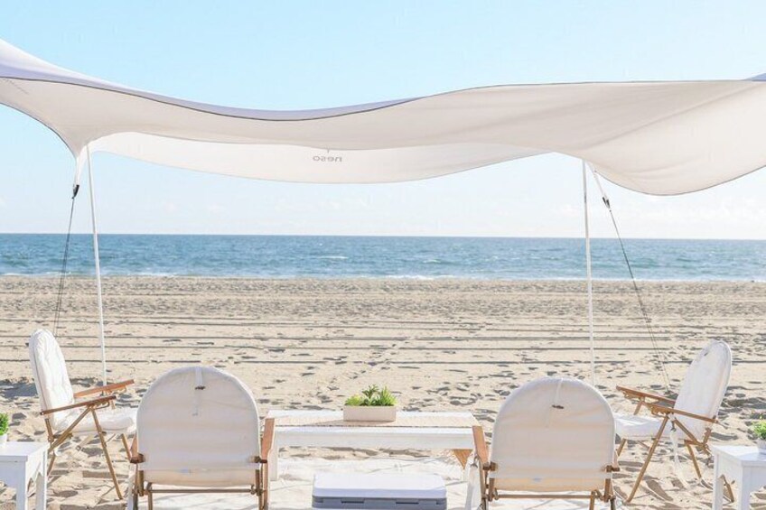 A beach with two white lounge chairs under a canopy, a table with a plant, and the ocean in the background. 2