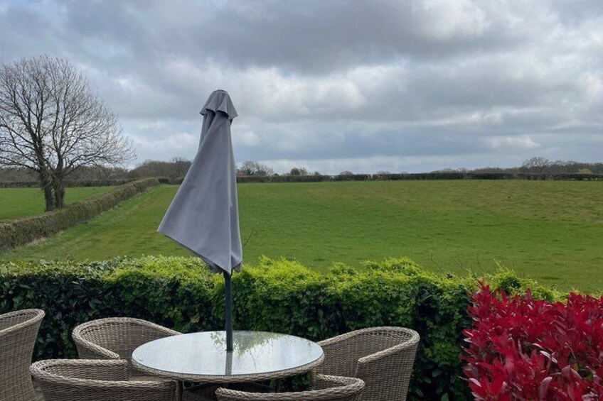 A round table with wicker chairs and a grey umbrella, set against a backdrop of a green field and a cloudy sky. 2