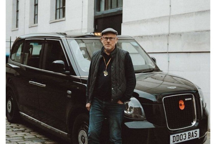 A man standing next to a black car with a license plate reading 'DD03 BAS' in an urban setting with buildings in the background. 4
