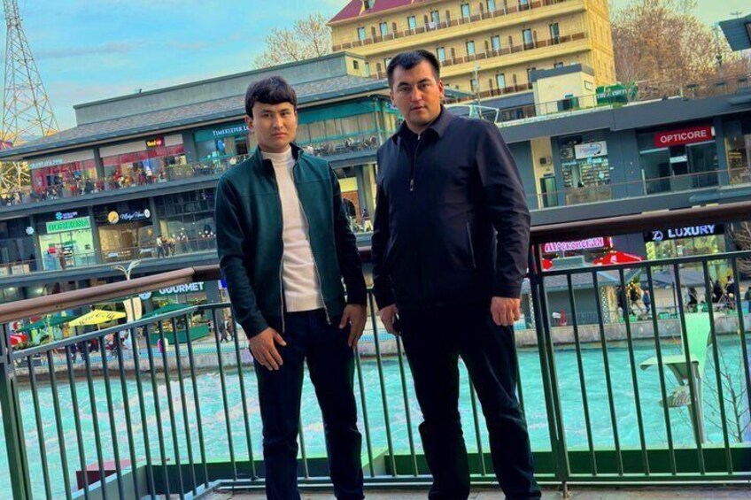 Two people standing on a balcony with a waterfront view, a building with shops in the background, and a railing in the foreground. 3