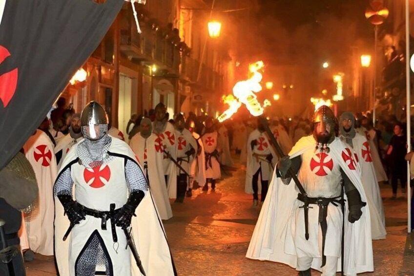 A medieval reenactment with participants in white and red costumes, carrying shields and swords, and a torch-lit street. 1
