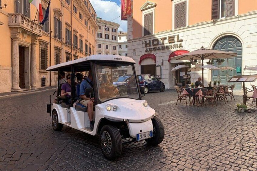 A white golf cart with passengers on a cobblestone street, a hotel, and outdoor seating in the background. 5
