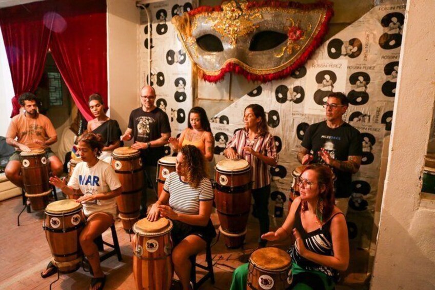 A group of people playing drums in a room with a decorative mask on the wall. 4
