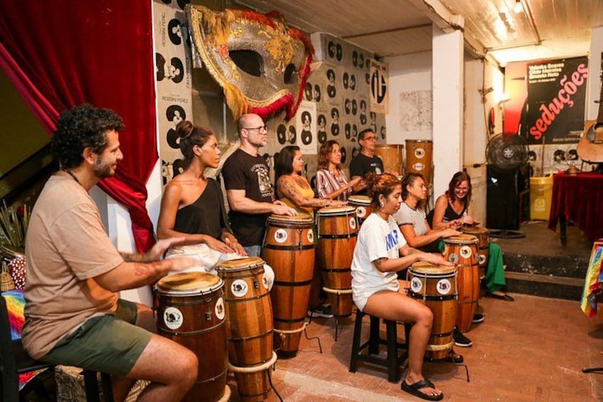 A group of people playing drums in a room with a red curtain and decorative masks on the wall. 2