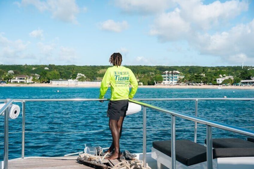 A person on a boat wearing a bright yellow jacket with 'Fiji' on it, standing on a deck with a railing, overlooking a body of water and a beach with buildings in the background. 3