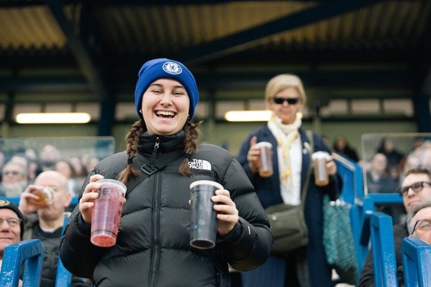 A person holding two cups, wearing a black jacket and a blue beanie, in a stadium with spectators. 4