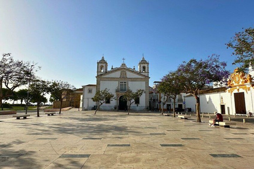 A plaza with a white church, benches, and trees. 5