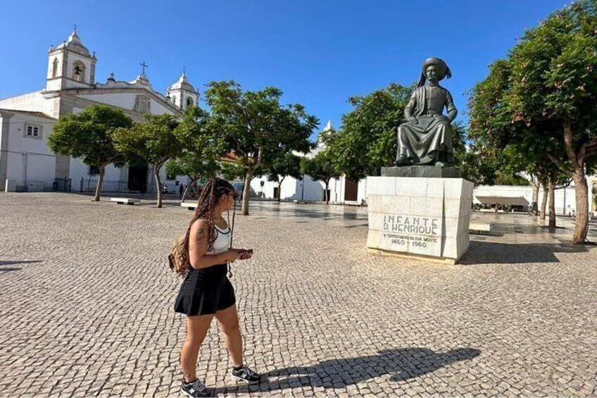 A statue of a historical figure in a square with cobblestone pavement, surrounded by trees and a church in the background. 2