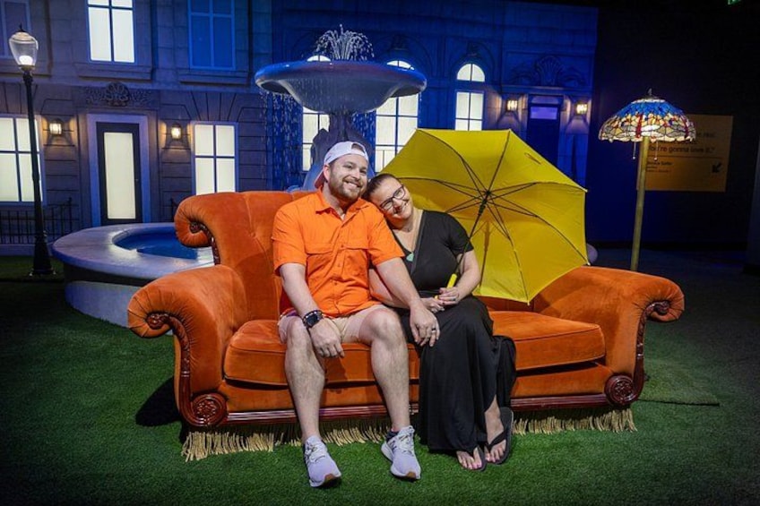 A couple sitting on an orange couch with a yellow umbrella in a courtyard with a fountain and a building in the background. 1