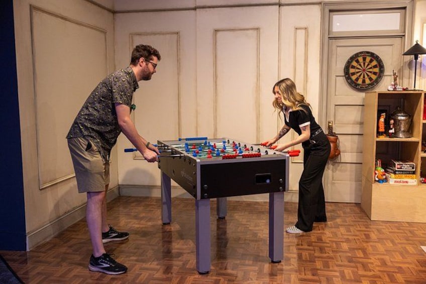 Two people playing foosball, a dartboard, and a shelf with various items. 2