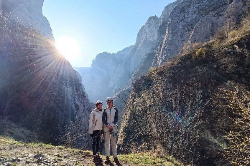 Two people standing in a canyon with a sun shining through. 1