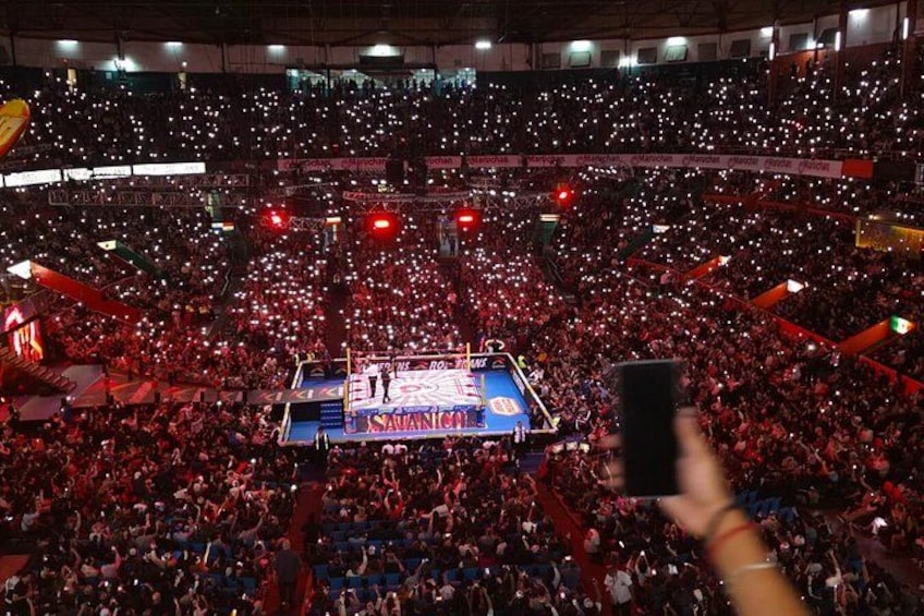 A large indoor arena filled with spectators, a stage with a blue backdrop, and a hand holding a smartphone. 2