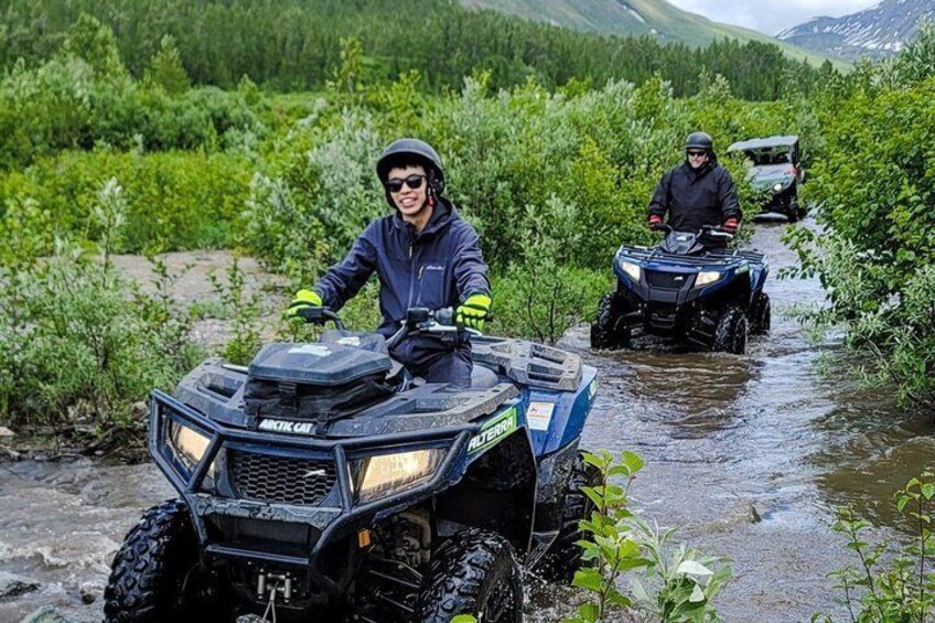 Two people on ATVs navigating a muddy stream in a forested area. 2