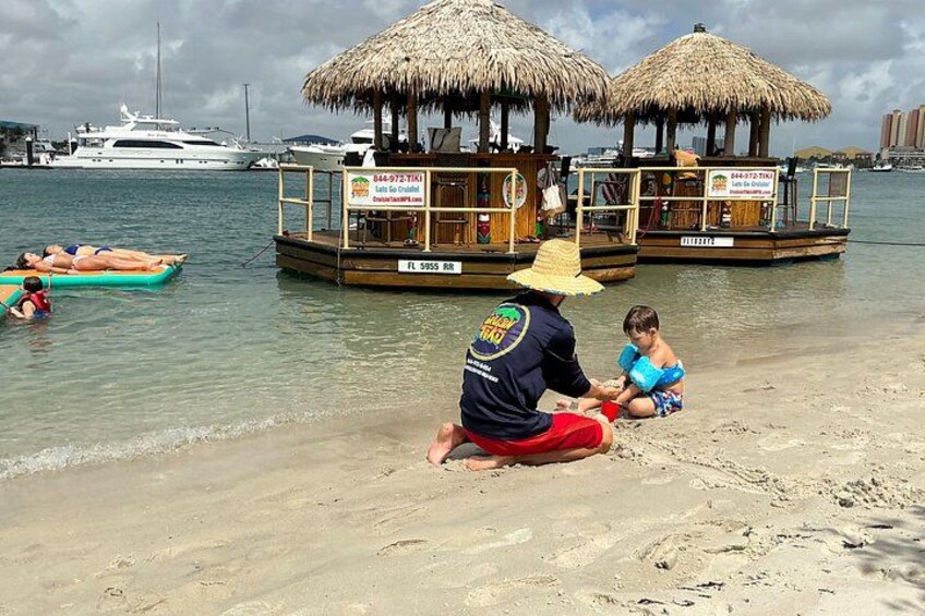 A beach with a man and a child playing in the sand, a thatched-roof structure, and a yacht in the background. 3