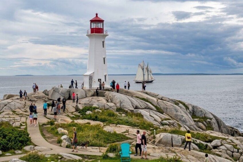 A lighthouse on a rocky outcrop with people walking around and a sailboat in the distance. 1
