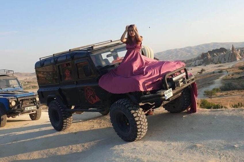 A person in a pink dress sitting on a black off-road vehicle with large tires, parked on a road with a rocky landscape in the background. 4