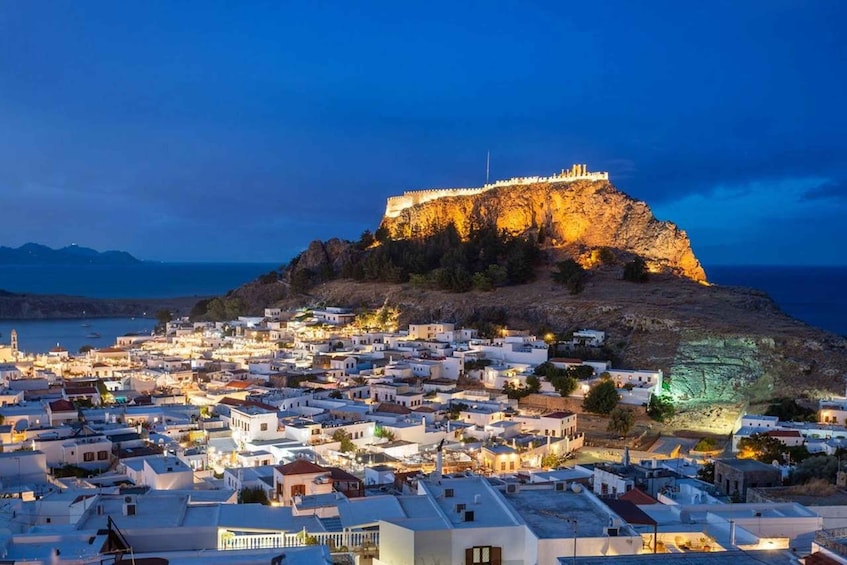 A coastal town at dusk with a prominent fortified hill in the background. 1