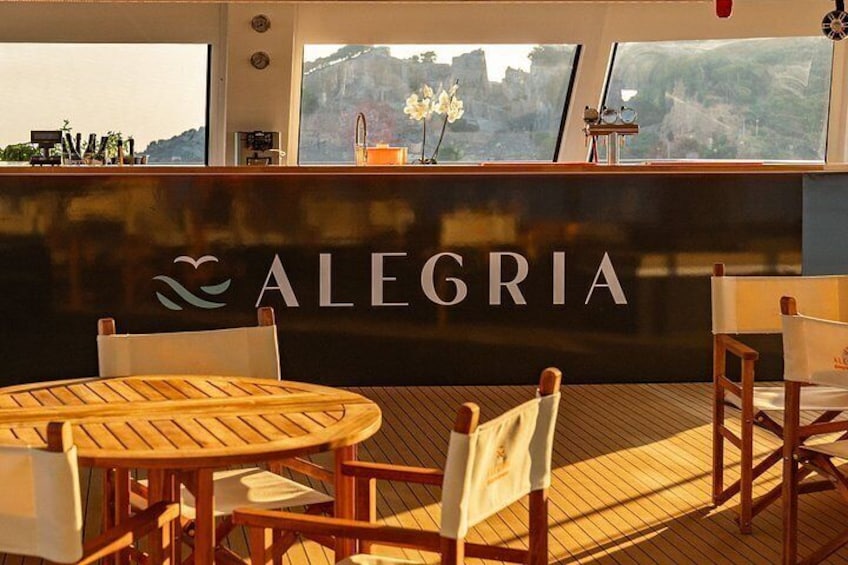 A wooden deck with a table and chairs, a bar area with the name 'ALEGRIA' on the wall, and a scenic view through the windows. 2