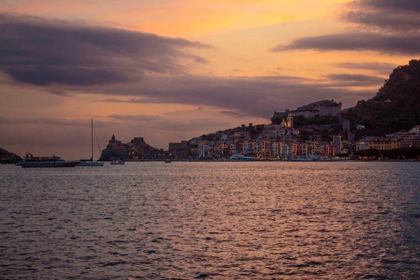 A coastal cityscape at sunset with buildings, a sailboat, and a hill with a fort. 2