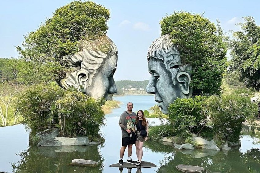 A couple standing in front of a sculpture with two heads, surrounded by water and greenery. 4