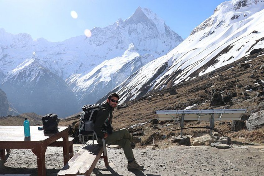 A person sitting on a bench with a backpack, a table with a water bottle, and a solar panel in a mountainous area. 5