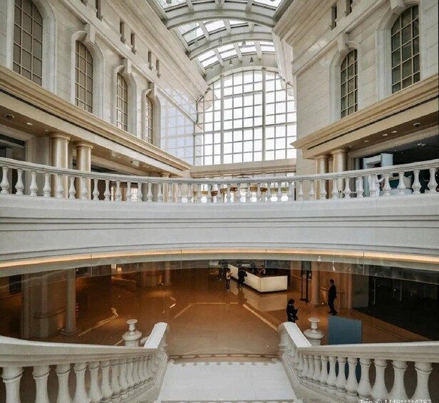A grand interior with a high ceiling and large windows, featuring a central open space with a reception desk and a lower level with a balcony. 3