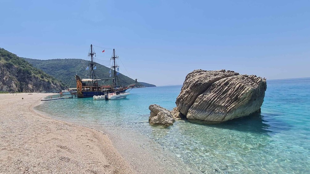 A clear beach with a large rock formation and a sailing ship anchored near the shore. 1