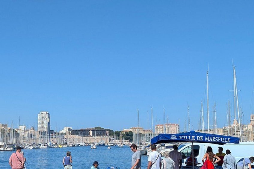 A marina with boats, a city skyline, and a blue tent with 'Ville de Marseille' written on it. 4