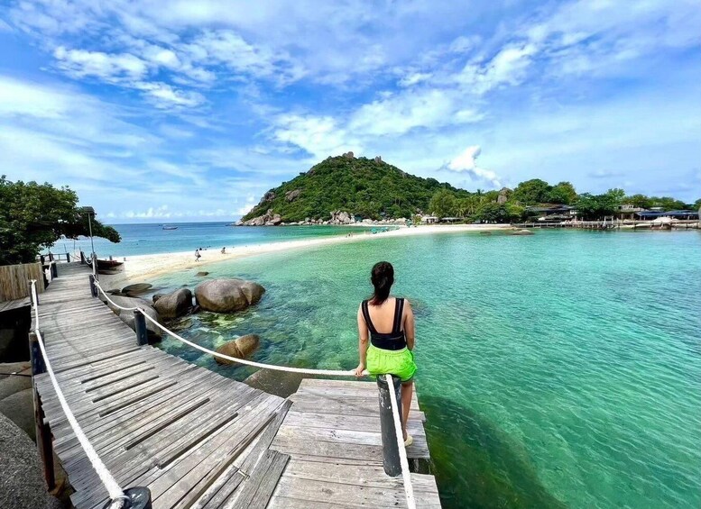A wooden pier leading into clear turquoise waters with a woman standing at the end, overlooking a beach and a hill with buildings in the distance. 5
