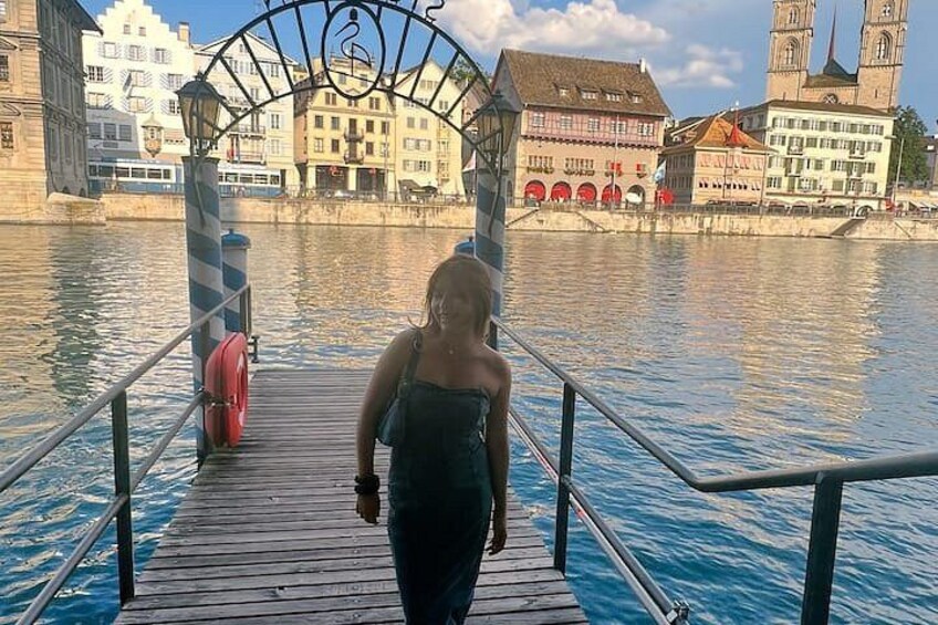 A person walking on a wooden pier with a metal railing, a red lifebuoy, and buildings in the background. 5