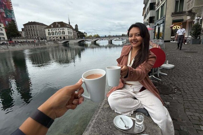 A person holding a cup of coffee, sitting by a river with buildings in the background. 2