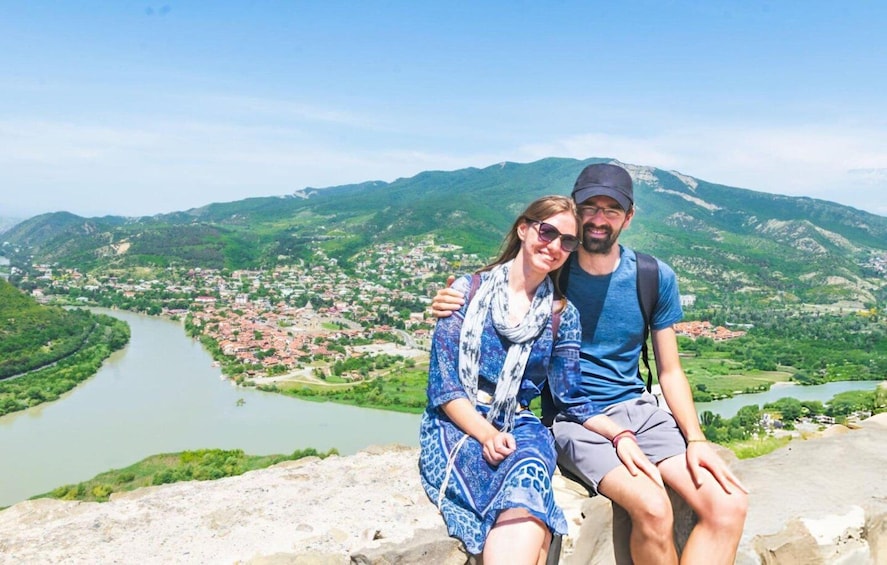 A couple sitting on a stone ledge overlooking a river and a town surrounded by mountains. 5