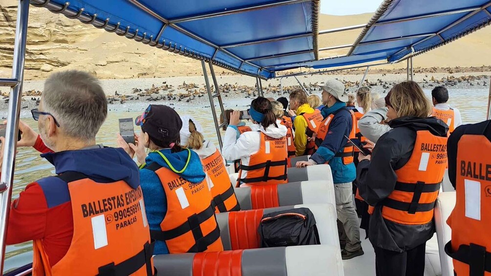 Un grupo de personas en un bote con chalecos salvavidas anaranjados con el texto 'BALLESTAS EXPEDICION PARACAS'. 4