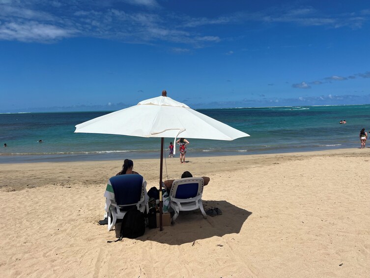 A beach with a white umbrella, two beach chairs, and a woman sitting. 5