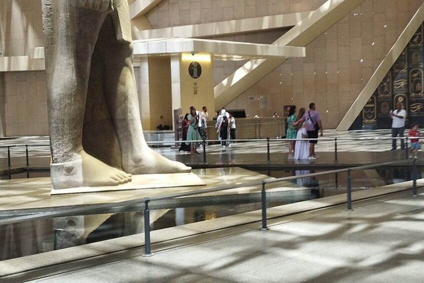 A large statue of a human figure in a museum with a reflective floor, visitors, and a modern architectural background. 4