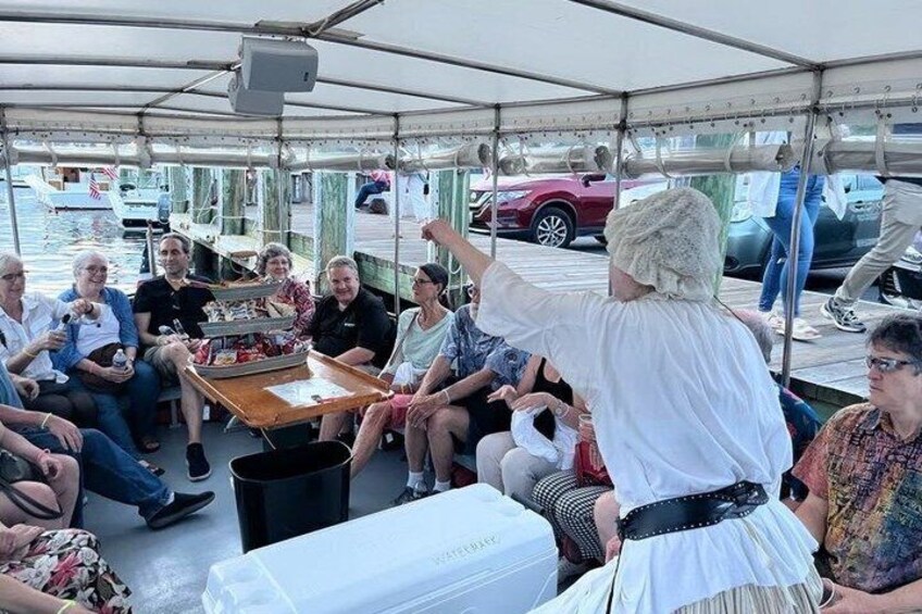 A group of people on a boat, some seated, some standing, under a covered area with a table and chairs. 1