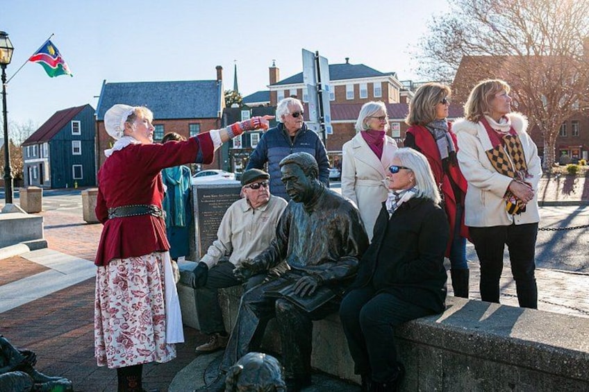 A historical reenactment scene with a person in period costume gesturing, a seated statue, and a group of onlookers. 4