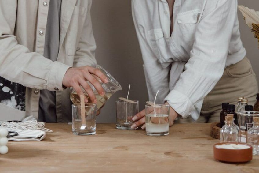 Two people pouring liquids into glasses on a wooden table with various bottles and a candle. 3