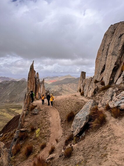 Een groep wandelaars op een rotsachtig bergpad, met schaarse begroeiing en bergen in de verte onder een bewolkte lucht. 2