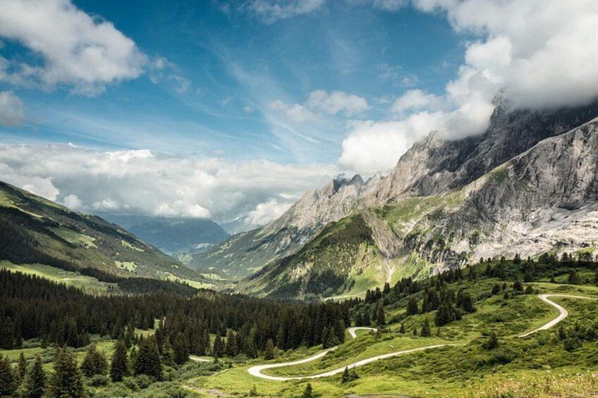 A mountainous landscape with a winding road, dense forests, and rocky cliffs under a partly cloudy sky. 4