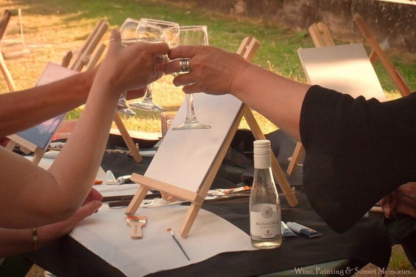 People toasting with wine glasses at an outdoor event with easels and a bottle of wine on the table. 5