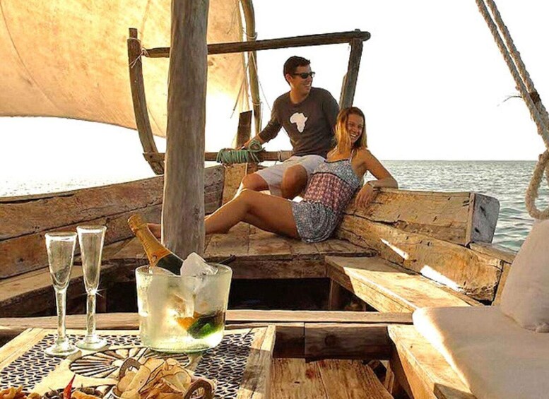 A couple on a wooden boat with a sail, sitting on a bench, and a table with drinks and snacks. 3