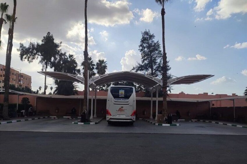 A bus station with a covered waiting area, palm trees, and a building in the background. 4