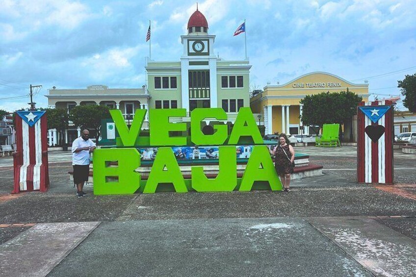 A sign with the words 'VEGA BAJA' in large letters, a clock tower, and a theater in the background. 4
